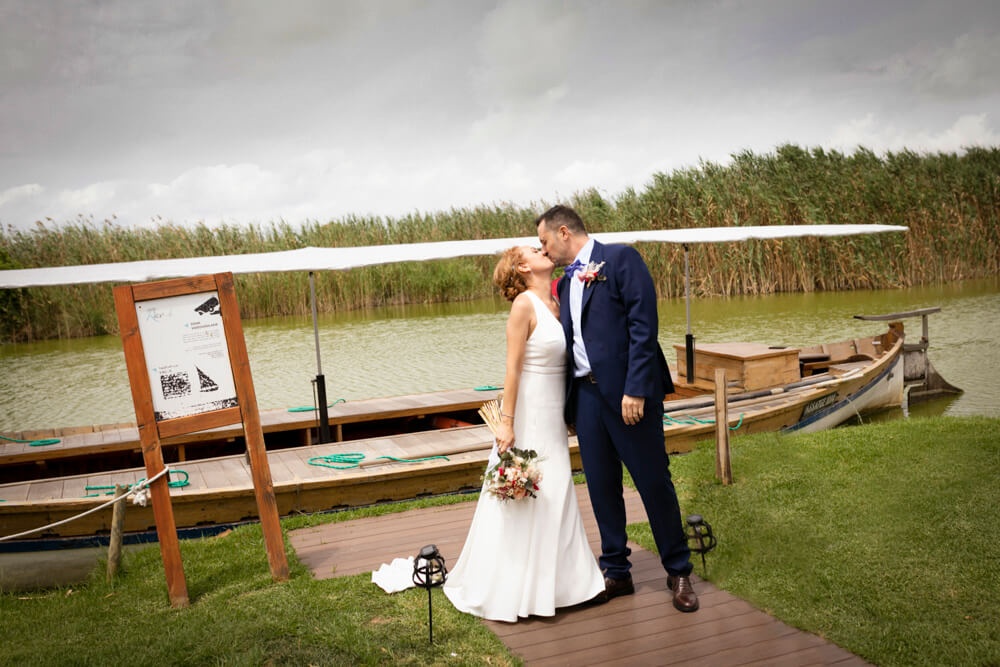 Pareja en su reportaje de boda, dando se un beso en Sesión de fotos de boda en el Nou Raco en Valencia. Están frente a una barca. Foto hecha por Mar Atienza