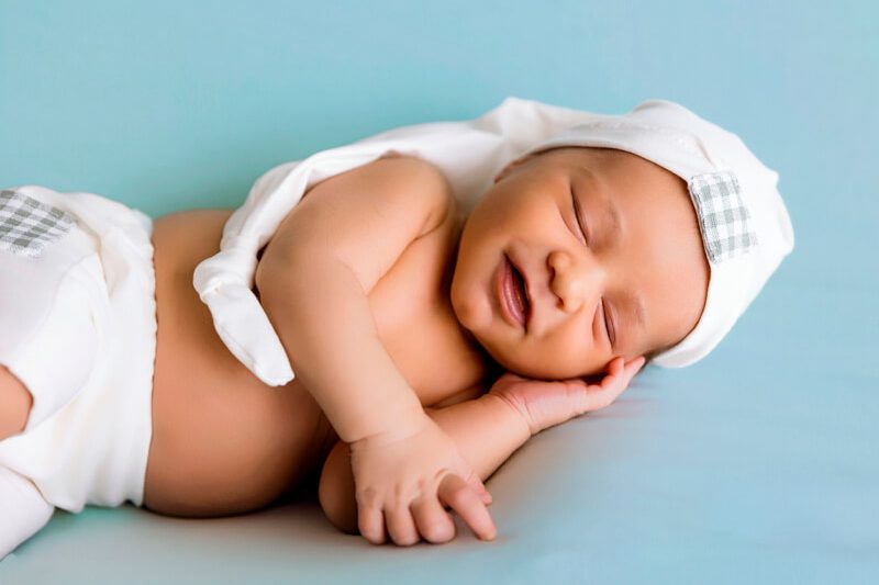 Bebé recién nacido sonriendo mientras duerme con gorro blanco en sesión de fotos