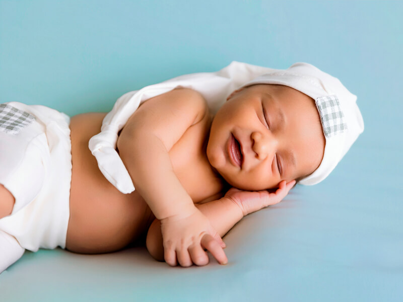 Bebé recién nacido sonriendo mientras duerme con gorro blanco en sesión de fotos