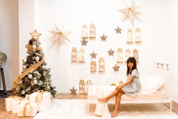 Niña en la acogedora habitación de El Pueblo Secreto de la Navidad, con una cama blanca, árbol navideño, regalos y pequeñas casitas iluminadas, en el estudio de Mar Atienza Fotografía en Valencia.